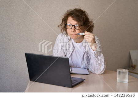 Young Woman Using Thermometer While Leaning Toward Laptop At Desk In Home Office With Notebook 142229580