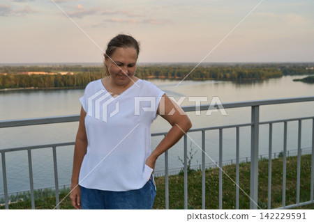 Adult Woman Standing By Railing With Hand On Hip And Looking Down Over River Landscape At Dusk Adult Woman Standing By Railing With Hand On Hip And Looking Down Over River Landscape At Dusk 142229591