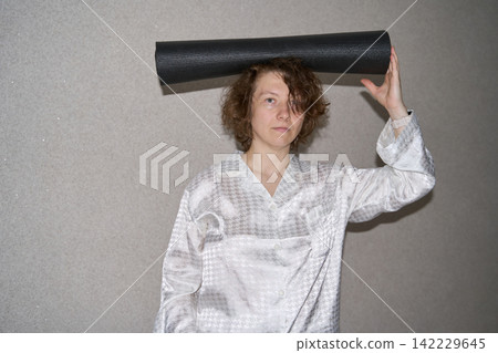 Woman Holding Rolled Black Exercise Mat Above Head While Standing Indoors, Playfully Balancing Woman Holding Rolled Black Exercise Mat Above Head While Standing Indoors, Playfully Balancing 142229645