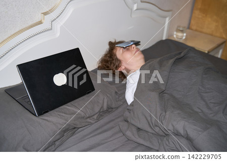 Woman Sleeping With Eye Mask Under Gray Blanket Beside Open Laptop In Bedroom, Showing Exhaustion Woman Sleeping With Eye Mask Under Gray Blanket Beside Open Laptop In Bedroom, Showing Exhaustion 142229705