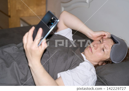 Woman Holding Smartphone Above Face While Reclining In Bed With Sleep Mask And Gray Blanket 142229727