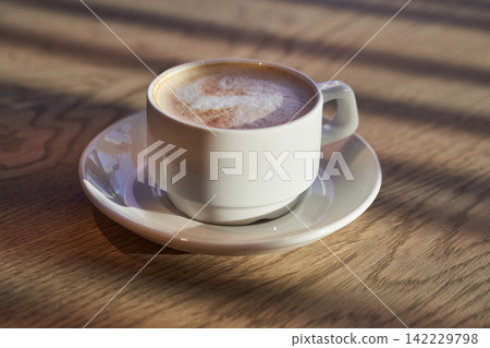 Cup Cappuccino On Saucer On Wooden Table With Morning Sunlight And Shadow Closeup Showing Foam Cup Cappuccino On Saucer On Wooden Table With Morning Sunlight And Shadow Closeup Showing Foam 142229798