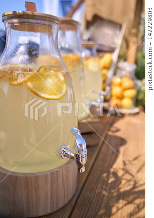 Glass Lemonade Dispensers With Citrus Slices Arranged On Rustic Wooden Table At Outdoor Summer Glass Lemonade Dispensers With Citrus Slices Arranged On Rustic Wooden Table At Outdoor Summer 142229803