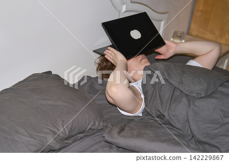 Woman Lying Under Gray Blanket With Open Laptop Covering Face In Bedroom, Expressing Digital Woman Lying Under Gray Blanket With Open Laptop Covering Face In Bedroom, Expressing Digital 142229867