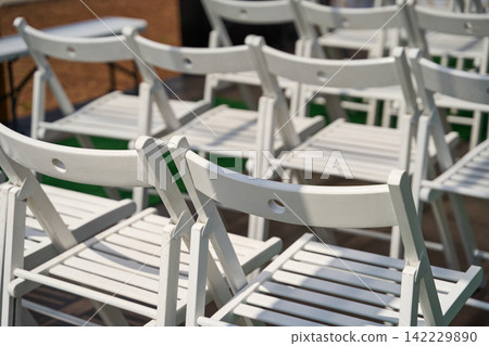 White Folding Chairs Clustered Together In Sunlit Outdoor Seating Area Before Ceremony On Warm Day 142229890