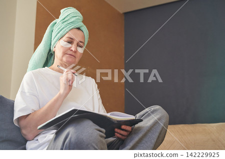Woman Reviewing Journal In Bedroom Wearing Towel Turban And Eye Patches While Seated Crosslegged Woman Reviewing Journal In Bedroom Wearing Towel Turban And Eye Patches While Seated Crosslegged 142229925