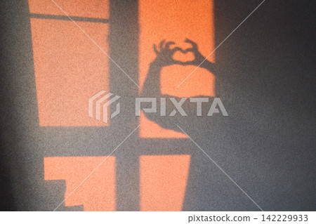 Shadow Hands Forming Heart Shape On Glowing Wall Beside Window Blinds At Sunset With Orange Light Shadow Hands Forming Heart Shape On Glowing Wall Beside Window Blinds At Sunset With Orange Light 142229933