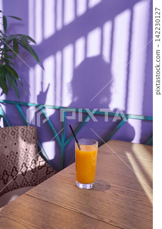 Glass Of Orange Juice Standing On Wooden Table Under Striped Shadow Of Person On Purple Wall Glass Of Orange Juice Standing On Wooden Table Under Striped Shadow Of Person On Purple Wall 142230127