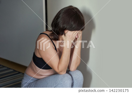 Adult Woman Covering Face While Seated On Low Step Beside Plain Wall In Small Apartment Interior Adult Woman Covering Face While Seated On Low Step Beside Plain Wall In Small Apartment Interior 142230175
