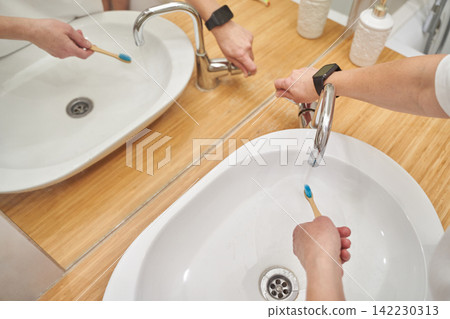 Adult Turning Faucet And Holding Toothbrush At Bathroom Sink With Wooden Countertop Mirror Adult Turning Faucet And Holding Toothbrush At Bathroom Sink With Wooden Countertop Mirror 142230313
