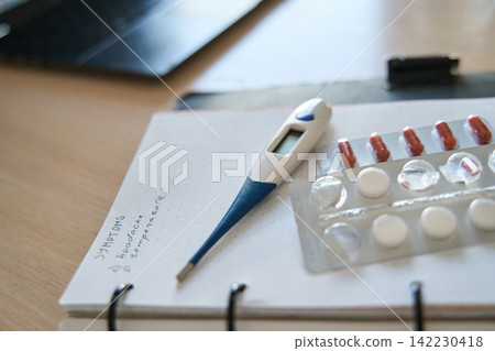 Close View Of Thermometer And Blister Pack On Notebook Beside Laptop In Bright Home Office Setting Close View Of Thermometer And Blister Pack On Notebook Beside Laptop In Bright Home Office Setting 142230418