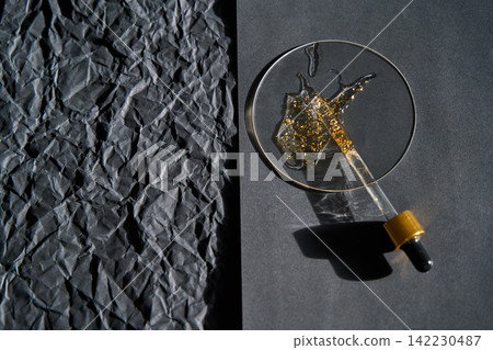 Dropper Resting With Spilled Gold Glitter Serum On Round Glass Dish Beside Crumpled Black Paper Dropper Resting With Spilled Gold Glitter Serum On Round Glass Dish Beside Crumpled Black Paper 142230487