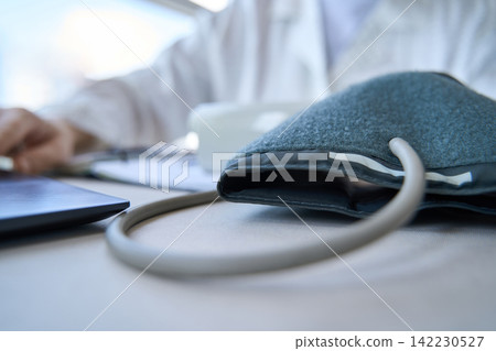 Blood Pressure Cuff Resting On Desk With Doctor In White Coat Blurred In Clinical Office Close Up Blood Pressure Cuff Resting On Desk With Doctor In White Coat Blurred In Clinical Office Close Up 142230527