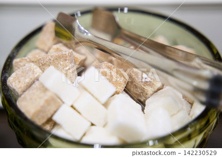 White And Brown Sugar Cubes Presented With Metal Tongs In Green Cut Glass Bowl On Counter, Closeup White And Brown Sugar Cubes Presented With Metal Tongs In Green Cut Glass Bowl On Counter, Closeup 142230529