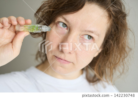 Woman Applying Rollon Under Eye In Neutral Indoor Setting Holding Small Glass Vial With Green Herbs Woman Applying Rollon Under Eye In Neutral Indoor Setting Holding Small Glass Vial With Green Herbs 142230748