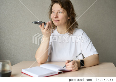 Woman Holding Smartphone For Voice Message While Writing In Open Notebook At Dining Table Woman Holding Smartphone For Voice Message While Writing In Open Notebook At Dining Table 142230933