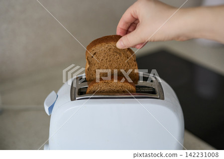 Hand Holding Single Brown Bread Slice Above White Two Slice Toaster On Kitchen Countertop Near Hand Holding Single Brown Bread Slice Above White Two Slice Toaster On Kitchen Countertop Near 142231008