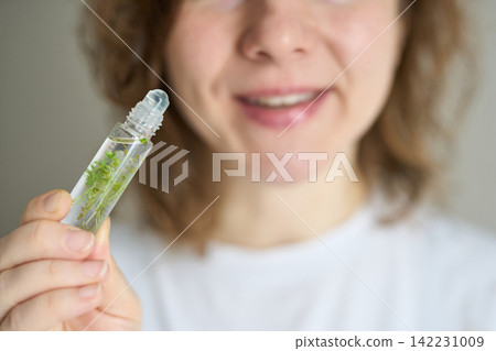 Woman Holding Glass Roller Bottle With Green Herbs Near Face Smiling In Neutral Indoor Closeup Woman Holding Glass Roller Bottle With Green Herbs Near Face Smiling In Neutral Indoor Closeup 142231009