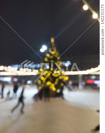 Tall Illuminated Christmas Tree With Bokeh Lights At Center Of Outdoor Ice Rink With Blurred Tall Illuminated Christmas Tree With Bokeh Lights At Center Of Outdoor Ice Rink With Blurred 142231173