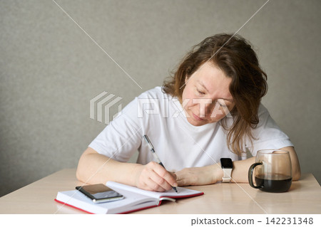 Woman Writing Notes Focused At Table With Open Notebook Smartphone And Pen Beside Mug Wearing White Woman Writing Notes Focused At Table With Open Notebook Smartphone And Pen Beside Mug Wearing White 142231348