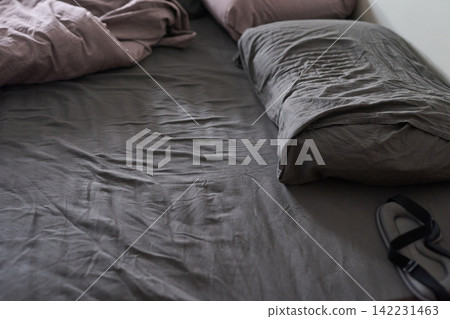 Unmade Bed With Wrinkled Grey Fitted Sheet Pillow And Discarded Slipper On Bedside In Natural Light Unmade Bed With Wrinkled Grey Fitted Sheet Pillow And Discarded Slipper On Bedside In Natural Light 142231463