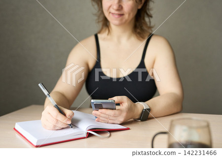 Woman Smiling While Jotting Notes In Notebook And Checking Smartphone At Wooden Table Indoors Woman Smiling While Jotting Notes In Notebook And Checking Smartphone At Wooden Table Indoors 142231466