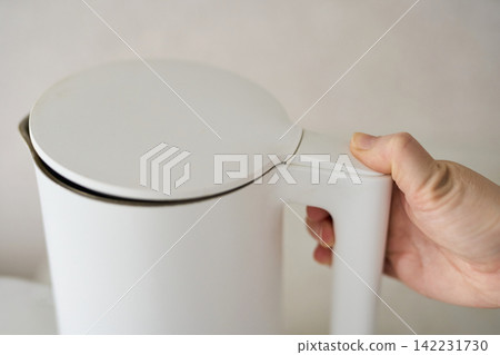 White Electric Kettle Lid Resting Closed While Hand Grips Handle On Countertop Closeup Showing Lid White Electric Kettle Lid Resting Closed While Hand Grips Handle On Countertop Closeup Showing Lid 142231730