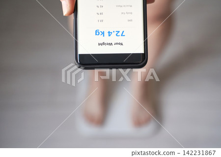 Overhead Close-Up Of Smartphone Held Above Blurred Feet On A Scale Showing 72.4 Kg For Fitness App Overhead Close-Up Of Smartphone Held Above Blurred Feet On A Scale Showing 72.4 Kg For Fitness App 142231867