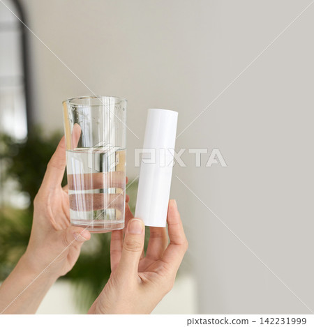 Hands Holding Glass Of Water And White Cylindrical Container In Bright Indoor Setting With Blurred Hands Holding Glass Of Water And White Cylindrical Container In Bright Indoor Setting With Blurred 142231999