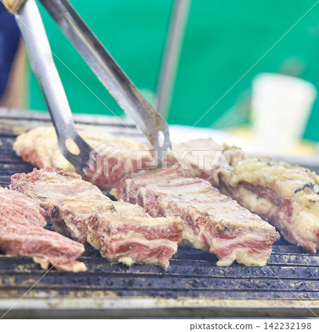 Pork Rib Slabs Being Flipped With Metal Tongs On Charcoal Grill At Outdoor Barbecue Market Stall Pork Rib Slabs Being Flipped With Metal Tongs On Charcoal Grill At Outdoor Barbecue Market Stall 142232198