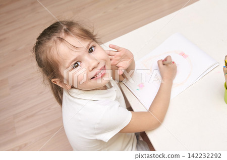 Caucasian young female child drawing with crayons on paper at home, smiling up Caucasian young female child drawing with crayons on paper at home, smiling up 142232292