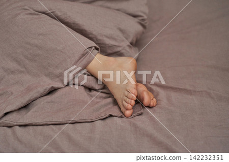 Female adult caucasian feet underneath soft brown bed cover in cozy morning relaxation scene 142232351