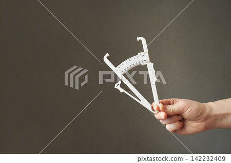 Caucasian adult hand holding caliper on grey background for body measurement and fitness assessment Caucasian adult hand holding caliper on grey background for body measurement and fitness assessment 142232409