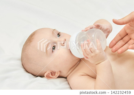 Caucasian Infant Drinking From Bottle With Adult Hand Offering Assistance On Soft White Surface Caucasian Infant Drinking From Bottle With Adult Hand Offering Assistance On Soft White Surface 142232459