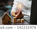 Hand holds glowing bulb above wooden house and blocks 142232978