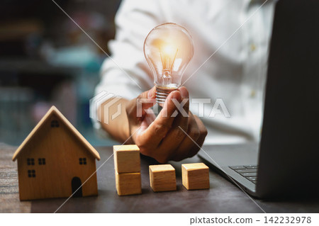 Hand holds glowing bulb above wooden house and blocks Hand holds glowing bulb above wooden house and blocks 142232978