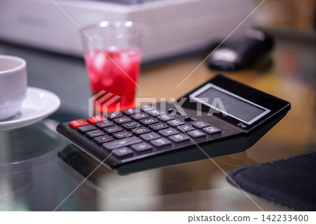 Black calculator glass office desk beside cup red drink representing accounting finance budgeting expenses business planning workplace calculations Black calculator glass office desk beside cup red drink representing accounting finance budgeting expenses business planning workplace calculations 142233400