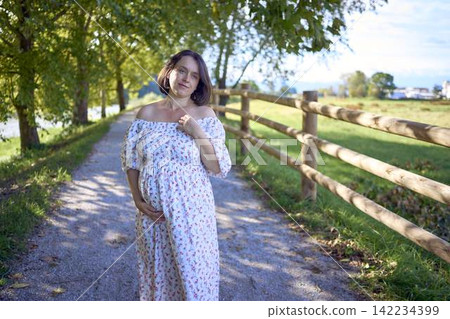 pregnant mature woman in floral dress walking on sunny rural pathway pregnant mature woman in floral dress walking on sunny rural pathway 142234399