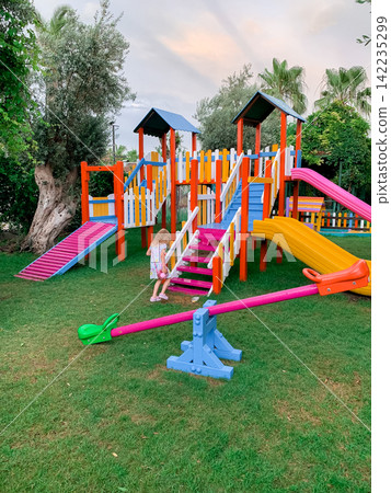 Young girl playing on a colorful playground with slides and seesaw in a green garden during summer evening Young girl playing on a colorful playground with slides and seesaw in a green garden during summer evening 142235299