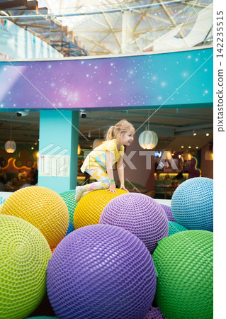 Little blonde girl in a yellow shirt playing on large colorful soft spheres in an indoor modern play area, child having fun and active leisure time in a bright entertainment center for kids Little blonde girl in a yellow shirt playing on large colorful soft spheres in an indoor modern play area, child having fun and active leisure time in a bright entertainment center for kids 142235515