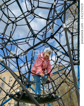 Active little girl wearing a pink jacket and a white hat climbing a tall rope web structure at an outdoor playground in the spring, looking down with a smile while playing on the climbing net Active little girl wearing a pink jacket and a white hat climbing a tall rope web structure at an outdoor playground in the spring, looking down with a smile while playing on the climbing net 142235629