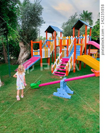 Young girl in a dress walking towards a colorful playground with slides and seesaw in a green garden Young girl in a dress walking towards a colorful playground with slides and seesaw in a green garden 142235858