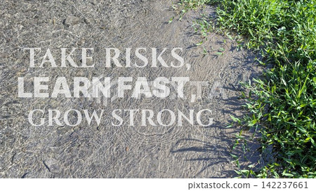A top down view shows inspirational motivational quote text stating "TAKE RISKS, LEARN FAST, GROW STRONG" overlaid on clear shallow river water flowing over sandy riverbed stones, lush green grass A top down view shows inspirational motivational quote text stating "TAKE RISKS, LEARN FAST, GROW STRONG" overlaid on clear shallow river water flowing over sandy riverbed stones, lush green grass 142237661