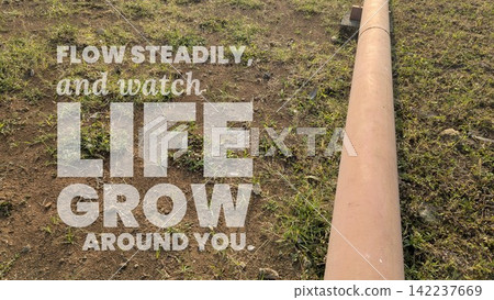 A high angle shot captures inspirational motivational quote text stating "FLOW STEADILY, watch LIFE GROW AROUND YOU." overlaid on a long pipe running across dry grassy ground, soil mixed with small A high angle shot captures inspirational motivational quote text stating "FLOW STEADILY, watch LIFE GROW AROUND YOU." overlaid on a long pipe running across dry grassy ground, soil mixed with small 142237669