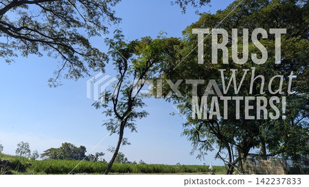 A horizontal wide angle shot captures inspirational motivational quote text stating "TRUST What MATTERS" overlaid on large lush green trees against a clear blue sky, grassy agricultural field 142237833