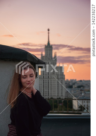 Woman poses on Moscow rooftop with Kotelnicheskaya Embankment Building and warm evening sky behind. Travel, urban lifestyle, city tourism, architecture, rooftop leisure, exploration. 142238217