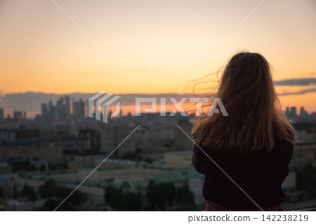 Woman watches sunset from Moscow rooftop with her back to camera above distant city skyline. Travel, urban lifestyle, contemplation, city exploration, rooftop leisure, tourism, freedom. 142238219