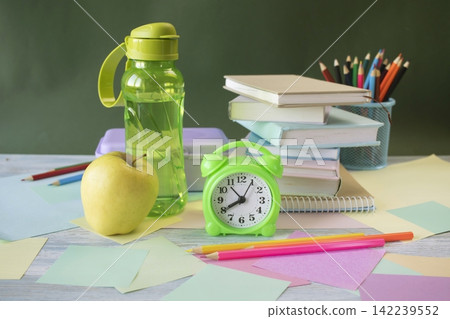 School supplies and healthy lunch on the study table. Bottle of water and lunch box and alarm clock, textbooks, colored pencils: back to school concept. School supplies and healthy lunch on the study table. Bottle of water and lunch box and alarm clock, textbooks, colored pencils: back to school concept. 142239552