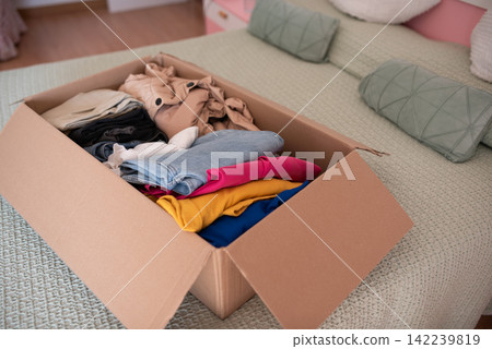 Cardboard box filled with folded clothes on a bed for moving Cardboard box filled with folded clothes on a bed for moving 142239819