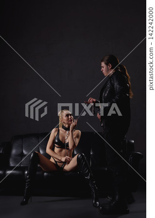 Man with chain leads woman in leather lingerie sitting on couch in dark room Man with chain leads woman in leather lingerie sitting on couch in dark room 142242000